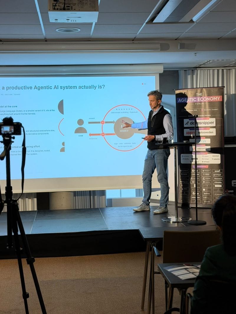 Bartosz Adam Gonczarek pointing at a slide that shows a user, the model at the core and the harness around it, which handles the prompt going in and the response coming out