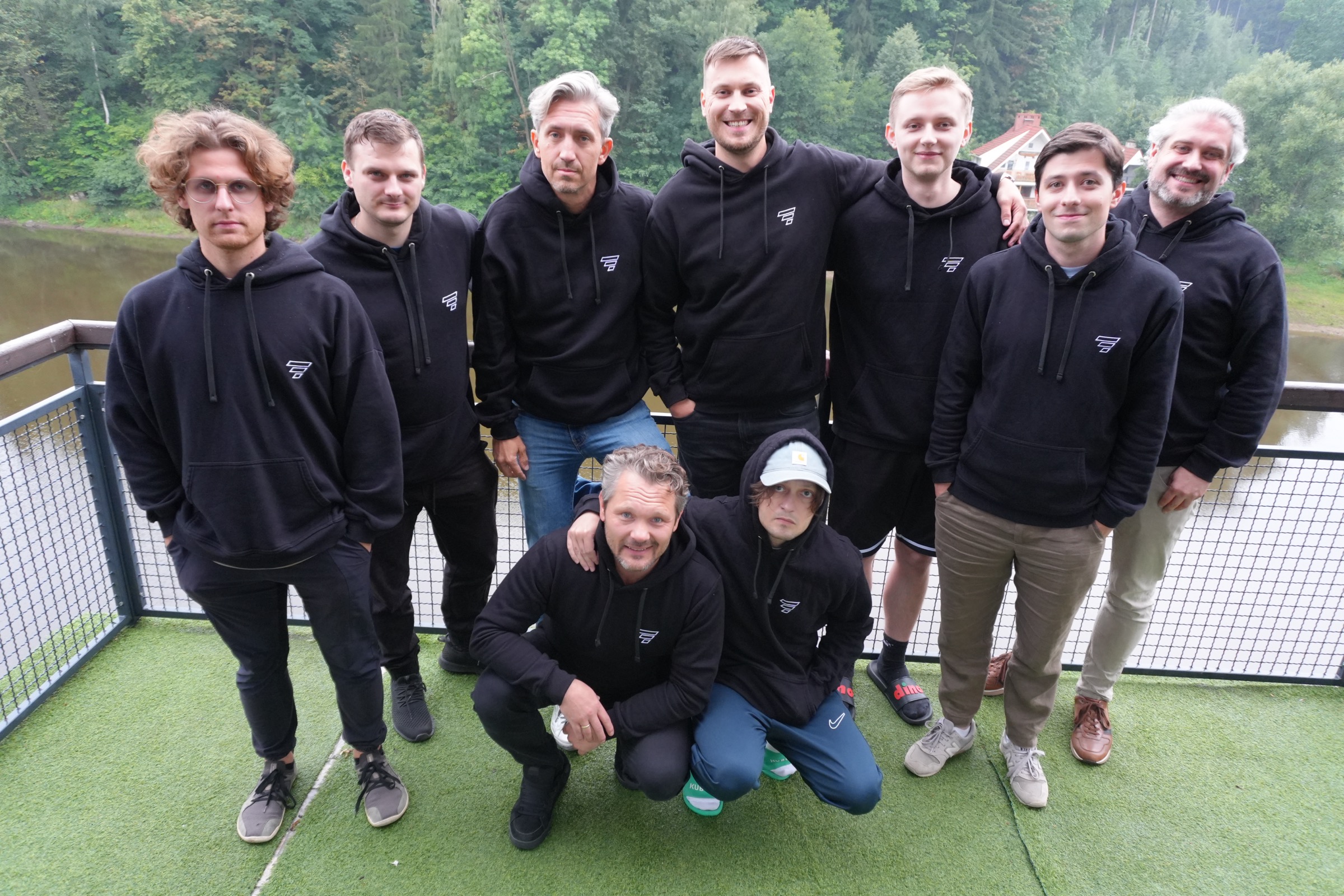 Nine members of the Vstorm core team in matching hoodies on a balcony overlooking forest and water at the August 2026 offsite